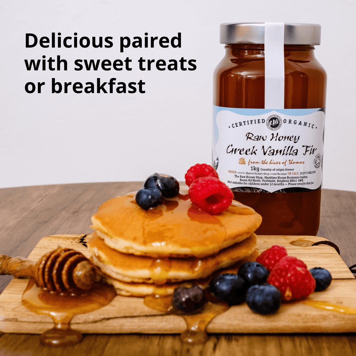 A jar of Certified Organic Raw Honey Greek Vanilla Fir syrup stands behind a stack of golden pancakes topped with blueberries and raspberries on a wooden board, with honey dripping around it and the text 'Delicious paired with sweet treats or breakfast' on a white background.