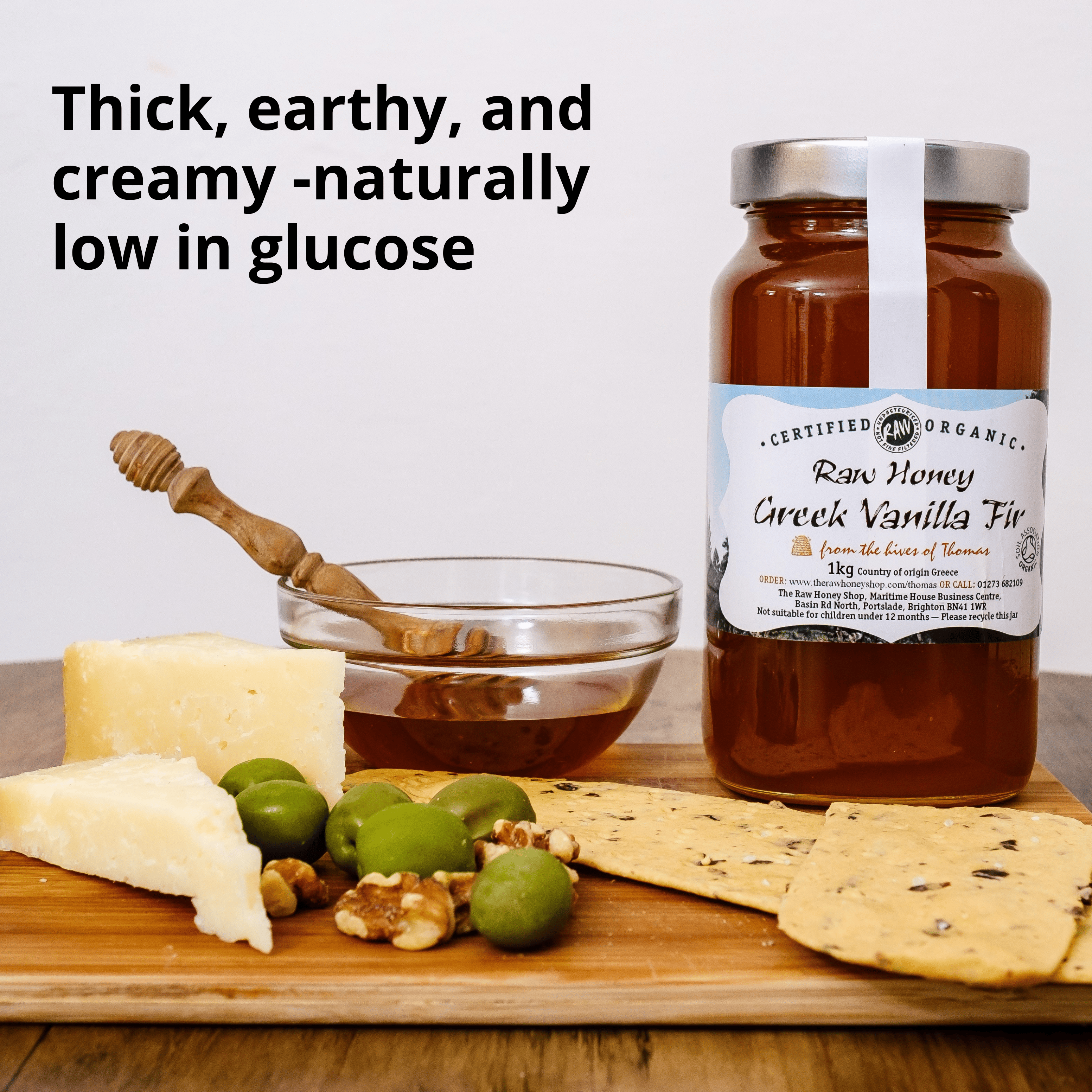 A jar of Raw Honey Creek Vanilla Fig honey displayed on a wooden board with cheese, olives, crackers, and a honey dipper. Text on the image reads 'Thick, earthy, and creamy -naturally low in glucose'.