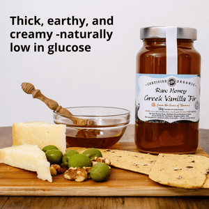 A jar of Raw Honey Creek Vanilla Fig honey displayed on a wooden board with cheese, olives, crackers, and a honey dipper. Text on the image reads 'Thick, earthy, and creamy -naturally low in glucose'.
