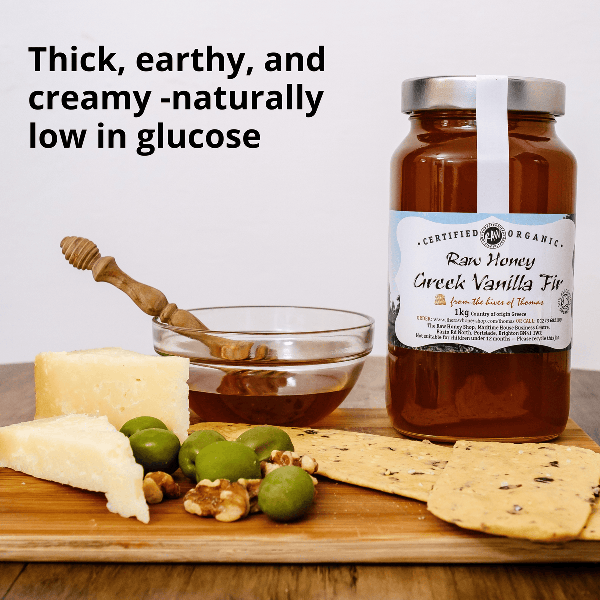 A jar of Raw Honey Creek Vanilla Fig honey displayed on a wooden board with cheese, olives, crackers, and a honey dipper. Text on the image reads 'Thick, earthy, and creamy -naturally low in glucose'.