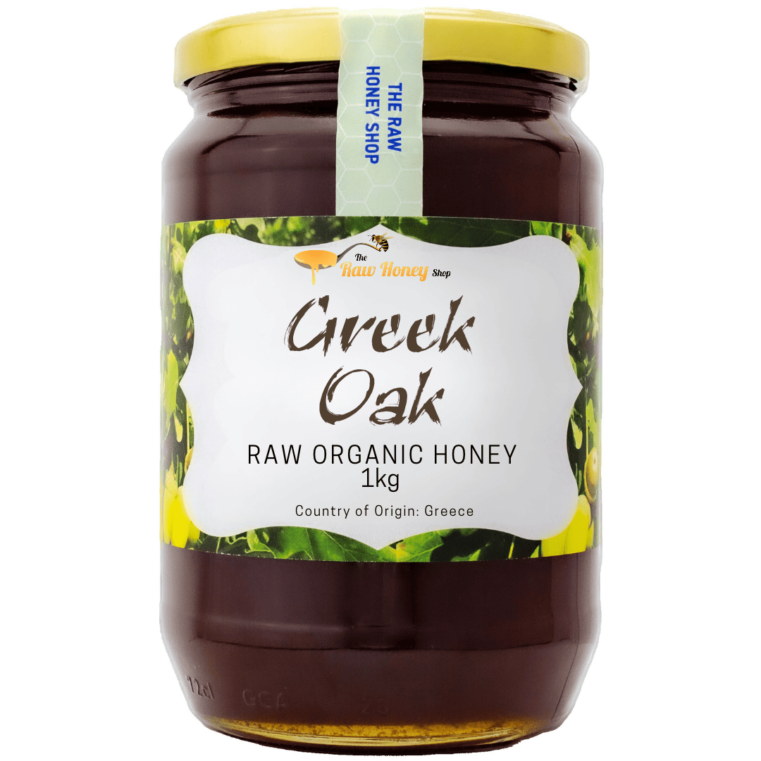A 1kg glass jar of Greek Oak Raw Organic Honey with a yellow lid and a white label featuring green and yellow leaf patterns, from The Raw Honey Shop.