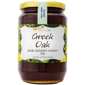 A 1kg glass jar of Greek Oak Raw Organic Honey with a yellow lid and a white label featuring green and yellow leaf patterns, from The Raw Honey Shop.