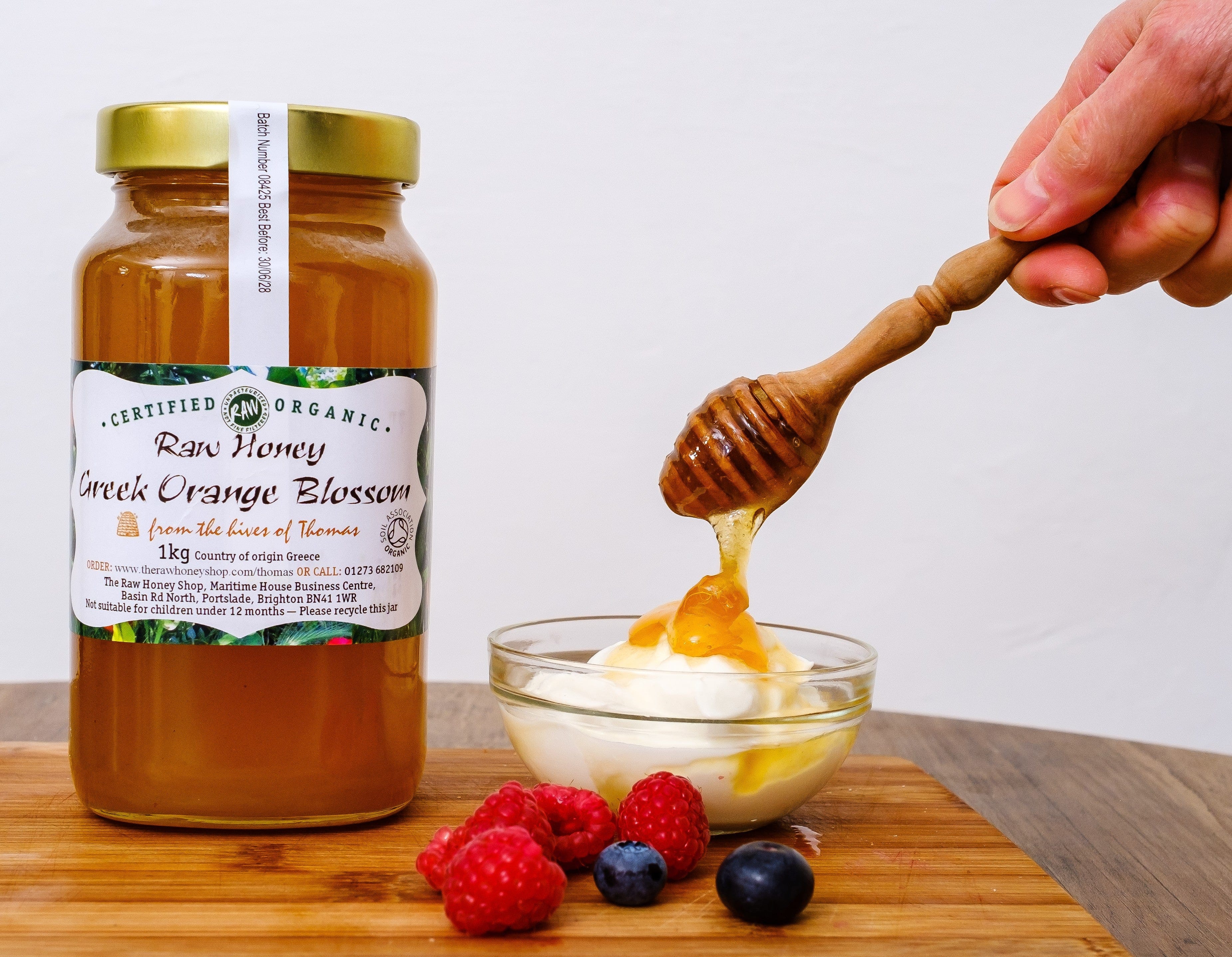 Bottle of raw honey with a wooden dipper over yogurt and fruit on a wooden board.