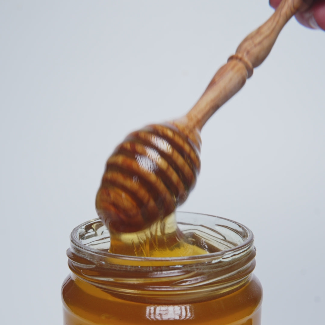 video of wooden honey dipper pouring into an open jar of  raw orange blossom honey.